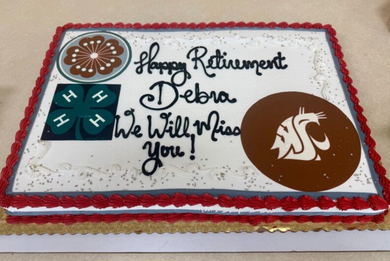 White sheet cake with crimson and gray edging, WSU Cougar Head logo and the 4-H and Master Gardener logos.  Text reads: "Happy Retirement, Debra. We Will Miss You!"