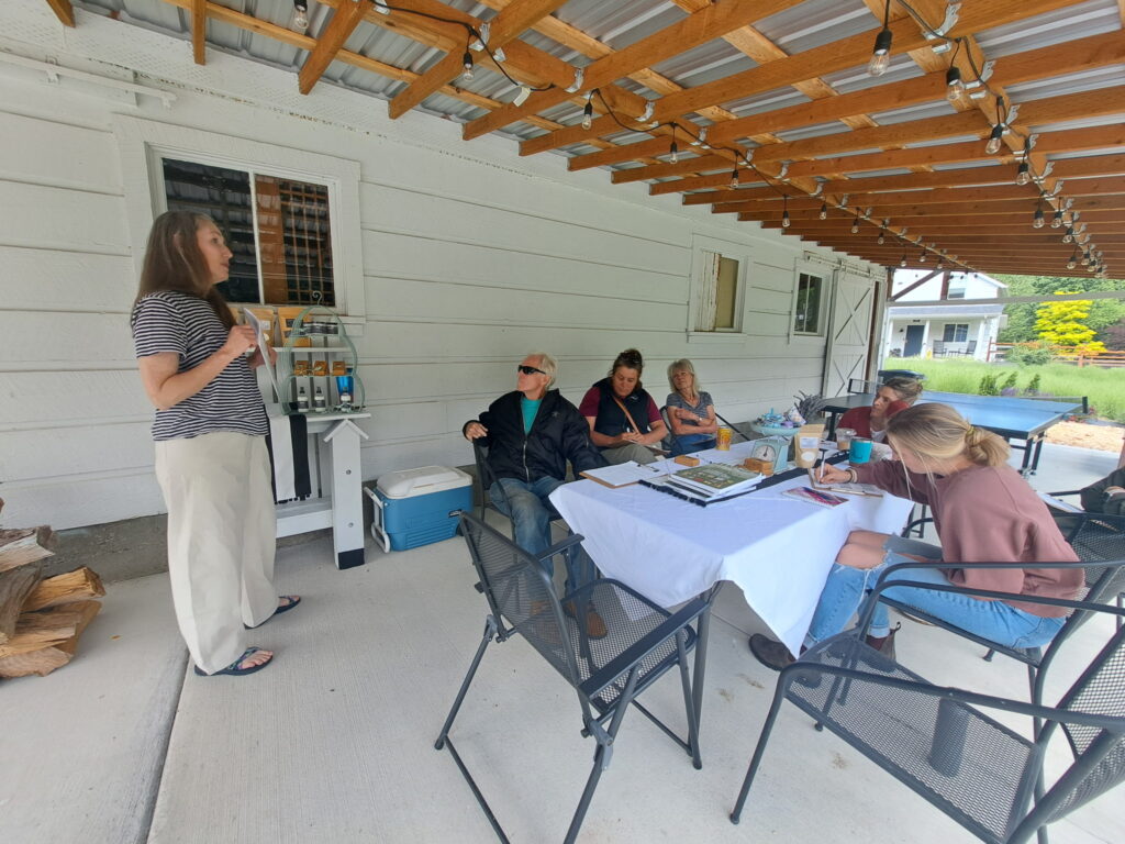 A woman stands talking while class participants are seated and taking notes; in an outdoor, covered seating area on the farm 
