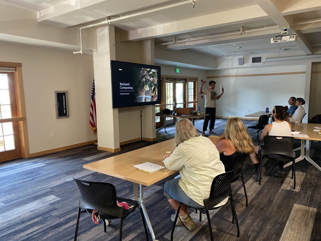A teacher stands in front of power point that says "Backyard Composting" in front of classroom of participants seated at long tables