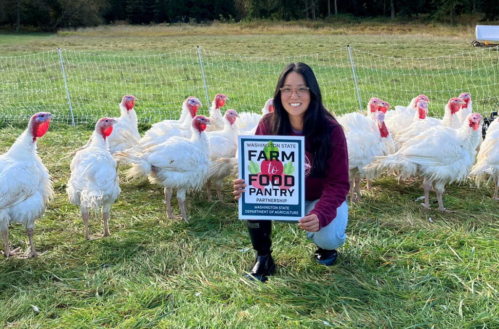 A woman holds a sign that says Farm to Food Pantry in a fenced pasture with several turkeys.