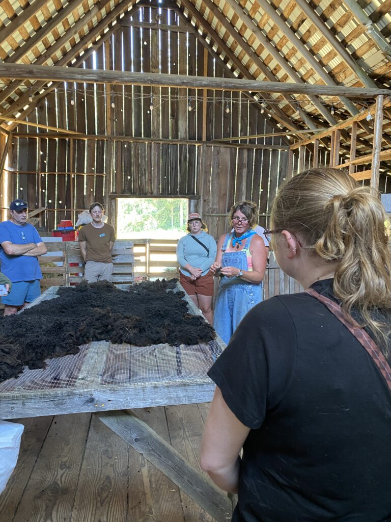 Participants stand around the skirting table, upon which rests a black fleece, and Roni talks about the skirting process