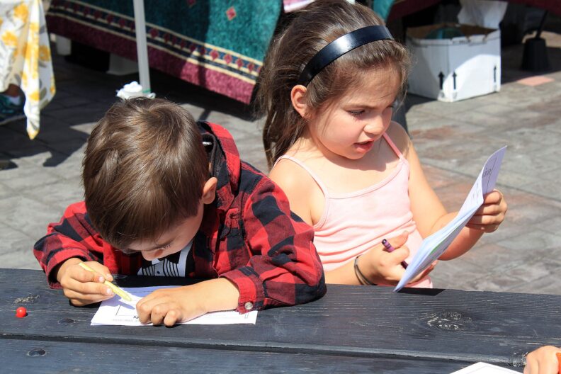 Two children sit at a picnic table, focused on drawing on sheets of paper with colorful crayons.