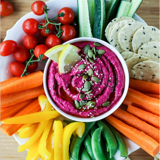 A bowl of beet hummus topped with seeds and lemon wedges, surrounded by various colorful veggies