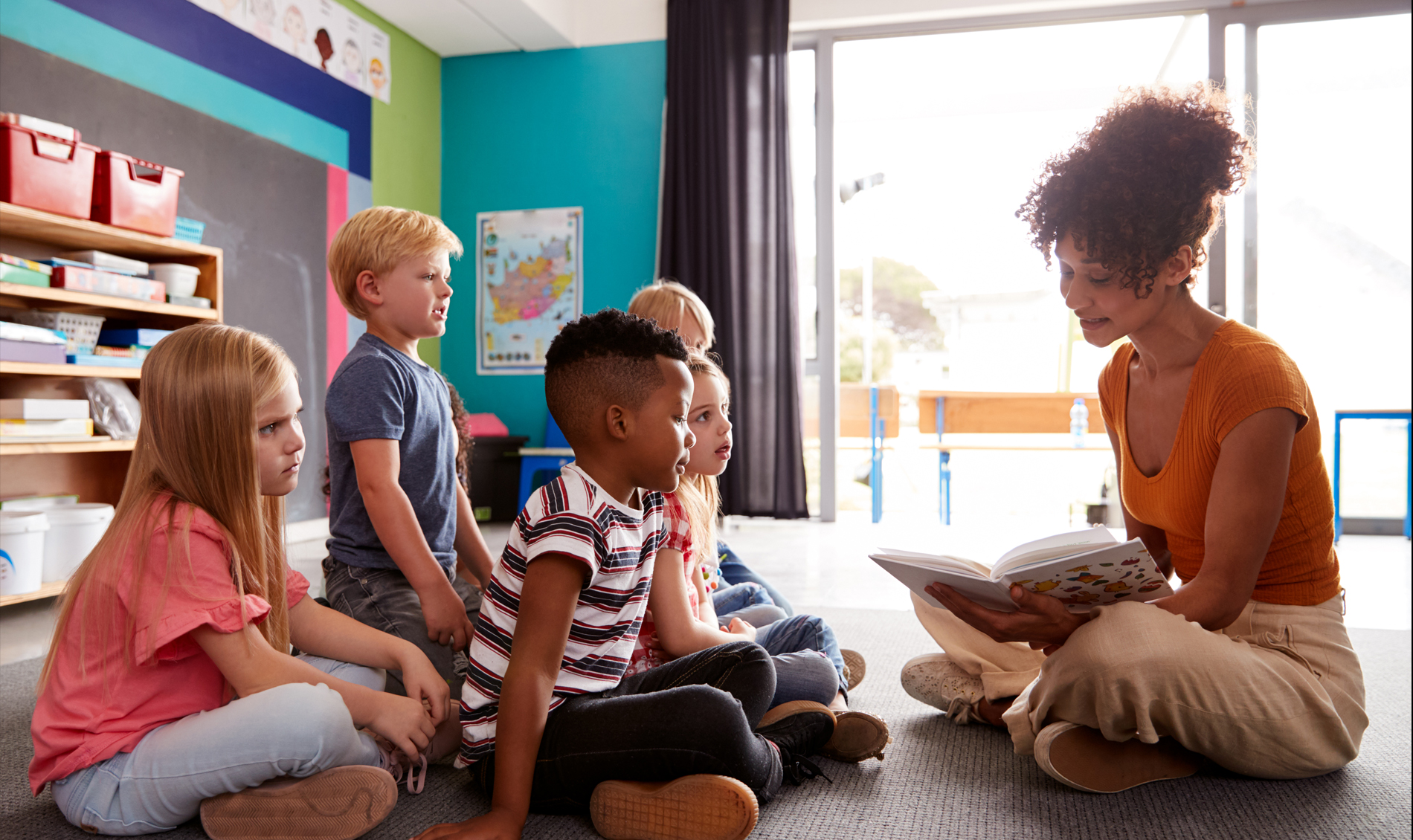 Teacher sits on a floor reading a book to four small students, also sitting on a floor.