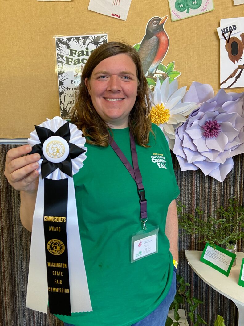 Woman in green t-shirt holding large ribbon at fairgrounds.