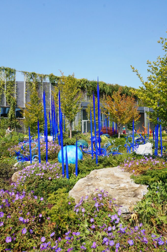 A glass-art exhibit surrounded by lush and flowering ground covers. Vines scale a building in the background.