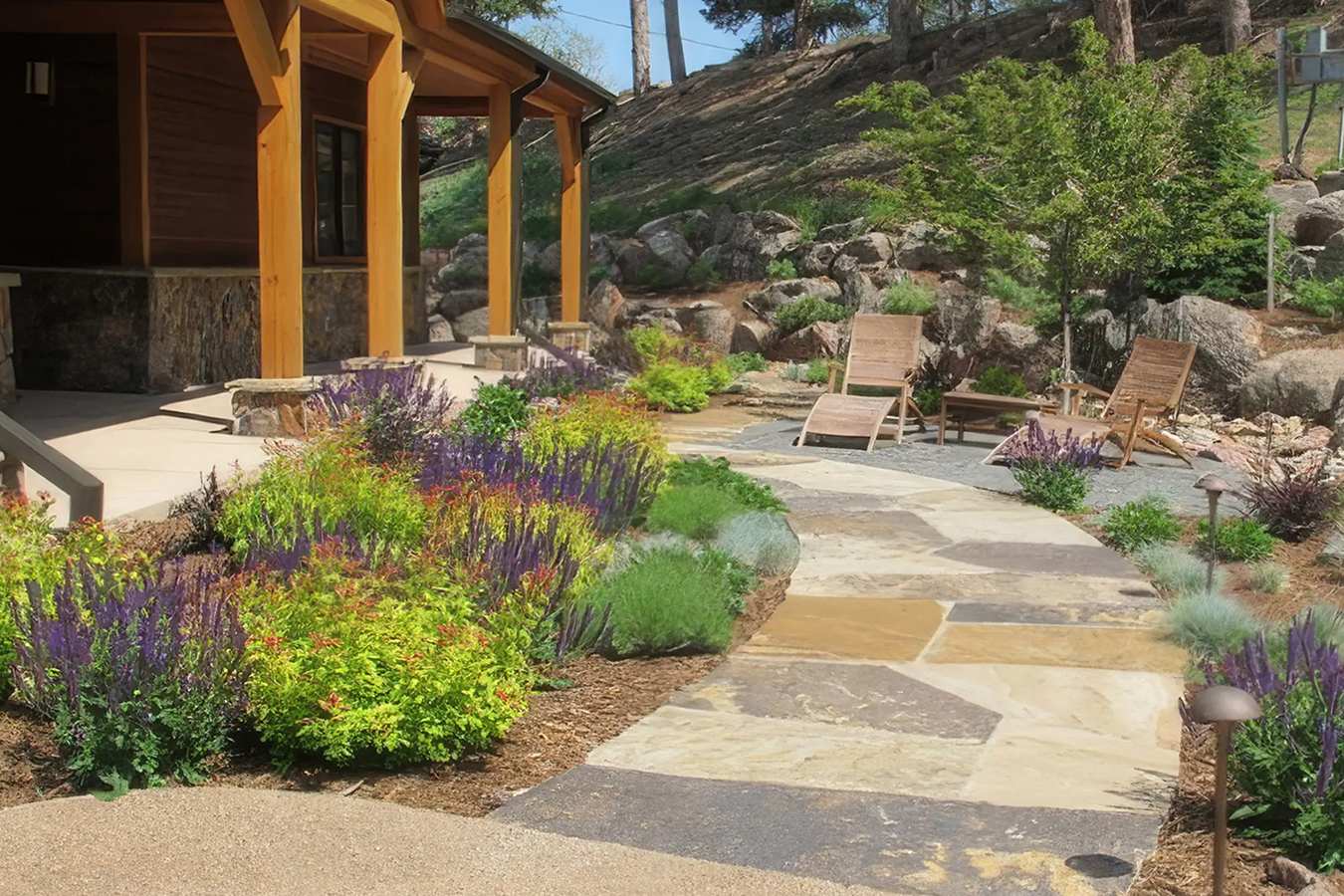 Dry landscape design with stone walkway, colorful drought-tolerant plants, and wooden lounge chairs near a house with a covered porch.
