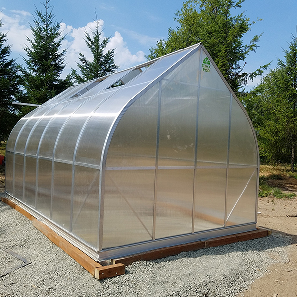 Small freestanding hobby greenhouse made of clear panels and metal framing, assembled on a gravel base with a wooden foundation.