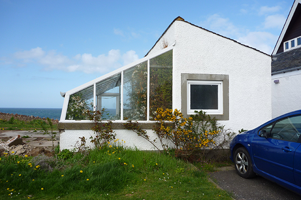 Attached greenhouse with slanted glass panels built against the side of a white house near the coast.