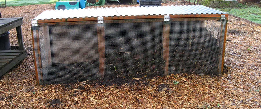 Screened outdoor composting unit with three different compartments and a corrugated metal roof.