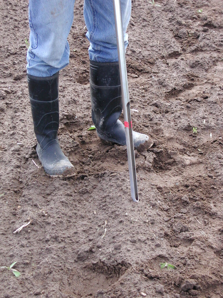 Man taking soil sample using a push probe. Someone handles a soil probe while wearing black rubber boots.