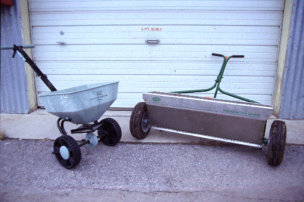 Two lawn spreaders, one small broadcast style and one larger drop style, sitting on pavement in front of a garage door.