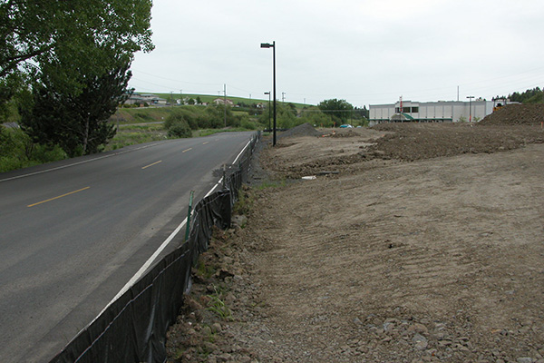 A short, black barrier alongside a road is staked upright, downhill from new construction.