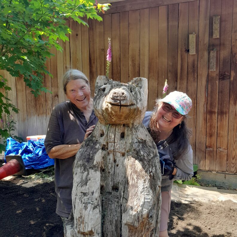 Master Gardener Marie Swanson and volunteer Joanne Tejeda pose smiling on either side of a wooden bear statue.