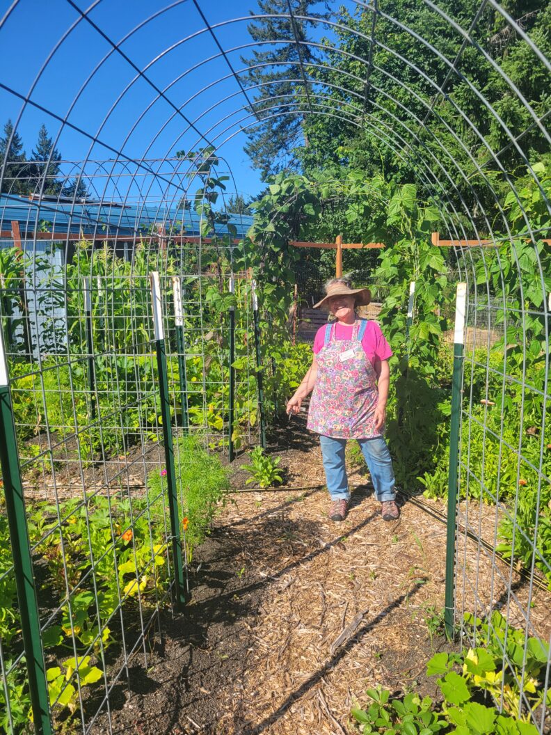 Master Gardener Amelia Savinova stands under trellis walkway in community garden.