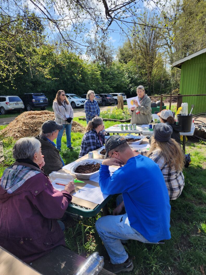 Six community garden workshops attendees sit around a picnic table at Catalyst Park while three Master Gardeners teach.