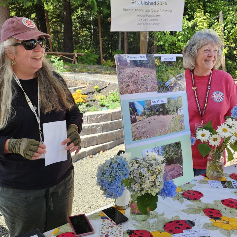 A photo of two people standing in front of a table with display board with photos. Person on right has long blond hair and is wearing black shirt and pants with red cap and person on left is wear red t-shirt and has short blond hair.