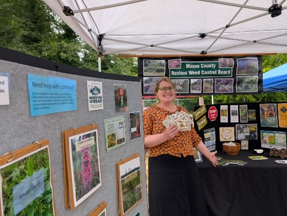 Person wearing black glasses, brown top and black pants is standing under a white canopy next to a large gray display panel with plant photos on it. Another large display is in the background behind the person.