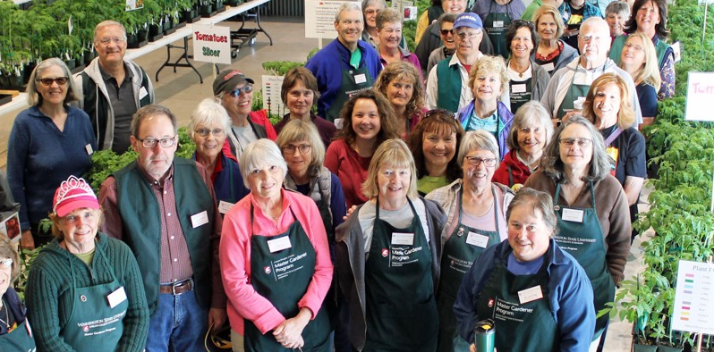 Large group of Master Gardener volunteers posing together at an indoor plant sale with seedlings on tables.