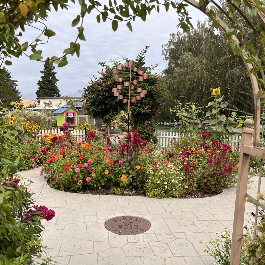 Colorful flower garden with circular planting bed, metal wind sculpture, and paved patio.