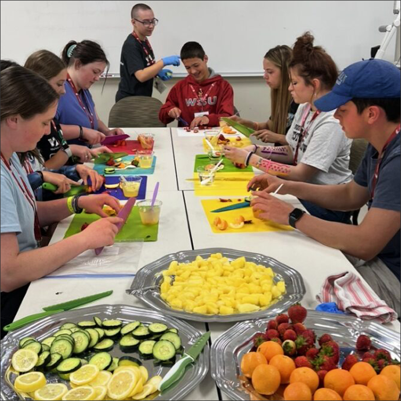 Image of multiple teens sitting around a table cutting fruit and vegetables