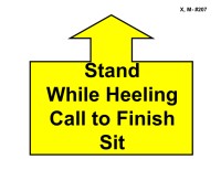 A large yellow arrow outlined in black points straight upward, attached to a wide rectangular base. Inside the base, bold black text reads “Stand While Heeling Call to Finish Sit.”