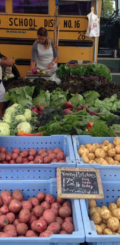 Potatoes and leafy vegetables for sale at a farmers market near a school bus.