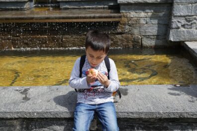 A young boy with a backpack sits on a stone ledge, intently examining an apple he holds in his hands.