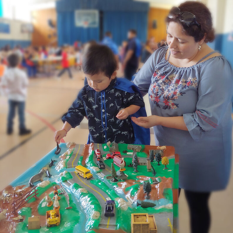 A young child interacts with a tabletop model of roads, rivers, and buildings with an adult.
