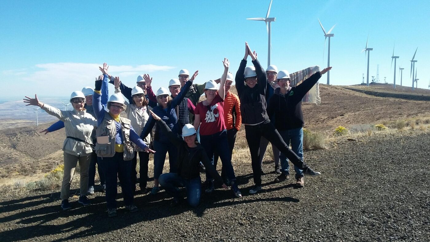 A group of people waving their hands and wearing hard hats while with a wind turbine farm in the background.