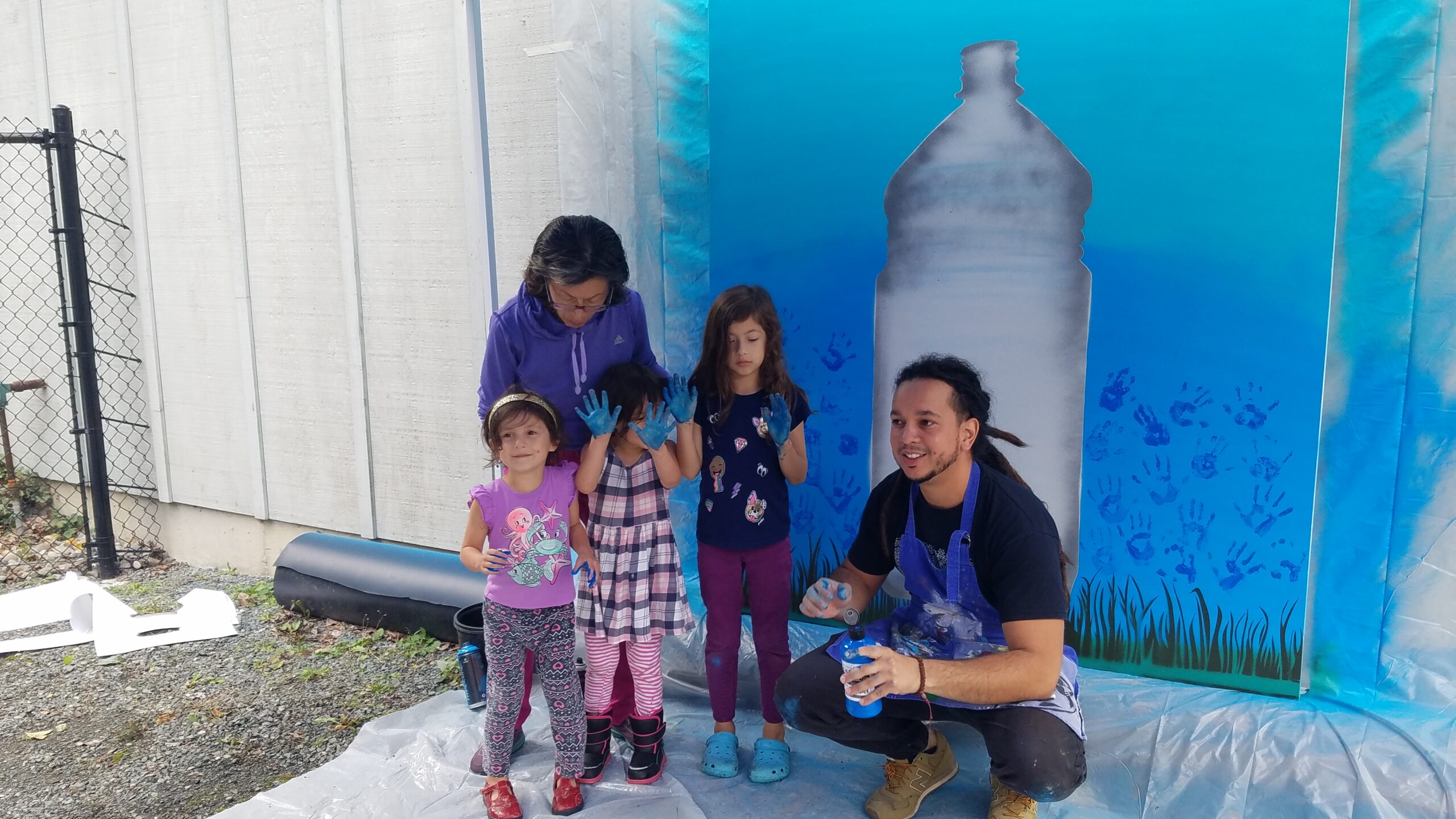 Adult and three children posing in front of a mural with a large painted water bottle.