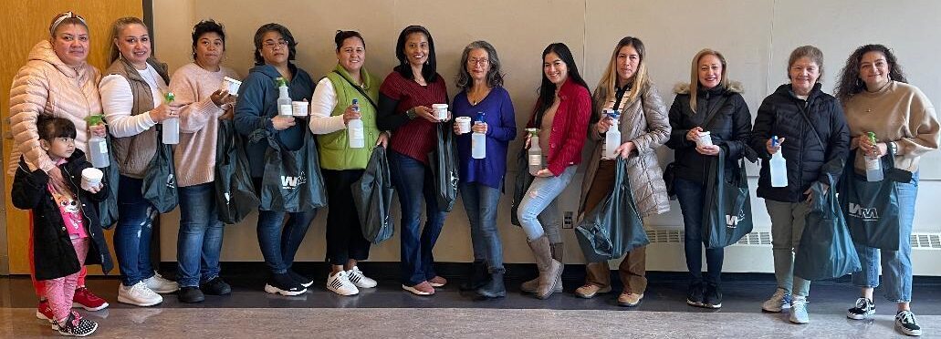 A group of twelve adults and one child stand in a row indoors, smiling and holding reusable bags and spray bottles.