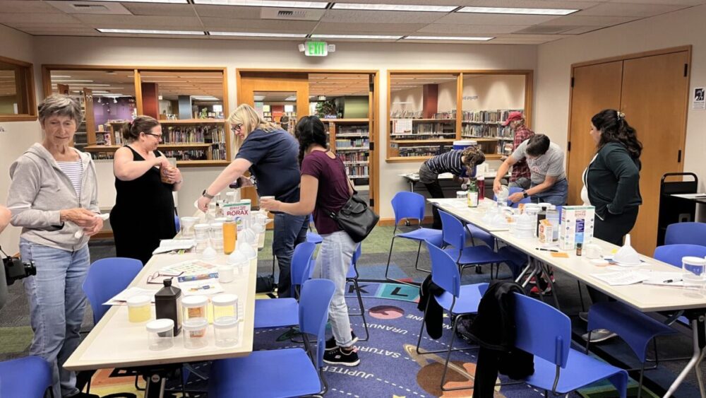Group of people surrounding two tables set with ingredients to make homemade cleaning supplies.
