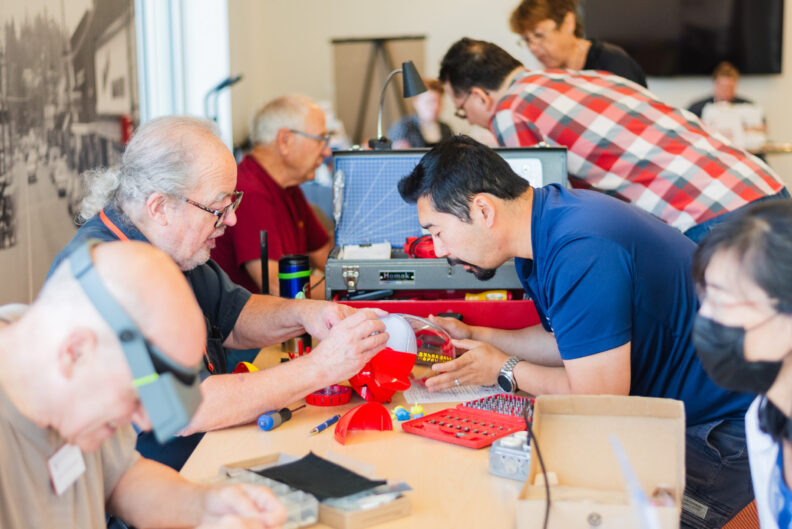 Six people on either side of a long table covered with tools, fixing objects and talking.