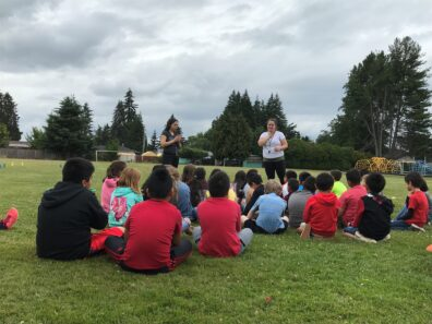 Children sit on grass listening to two instructors speaking outdoors.