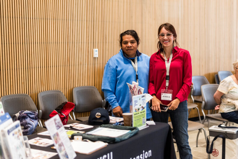 Two volunteers standing and smiling by the Repair Cafe check-in table.