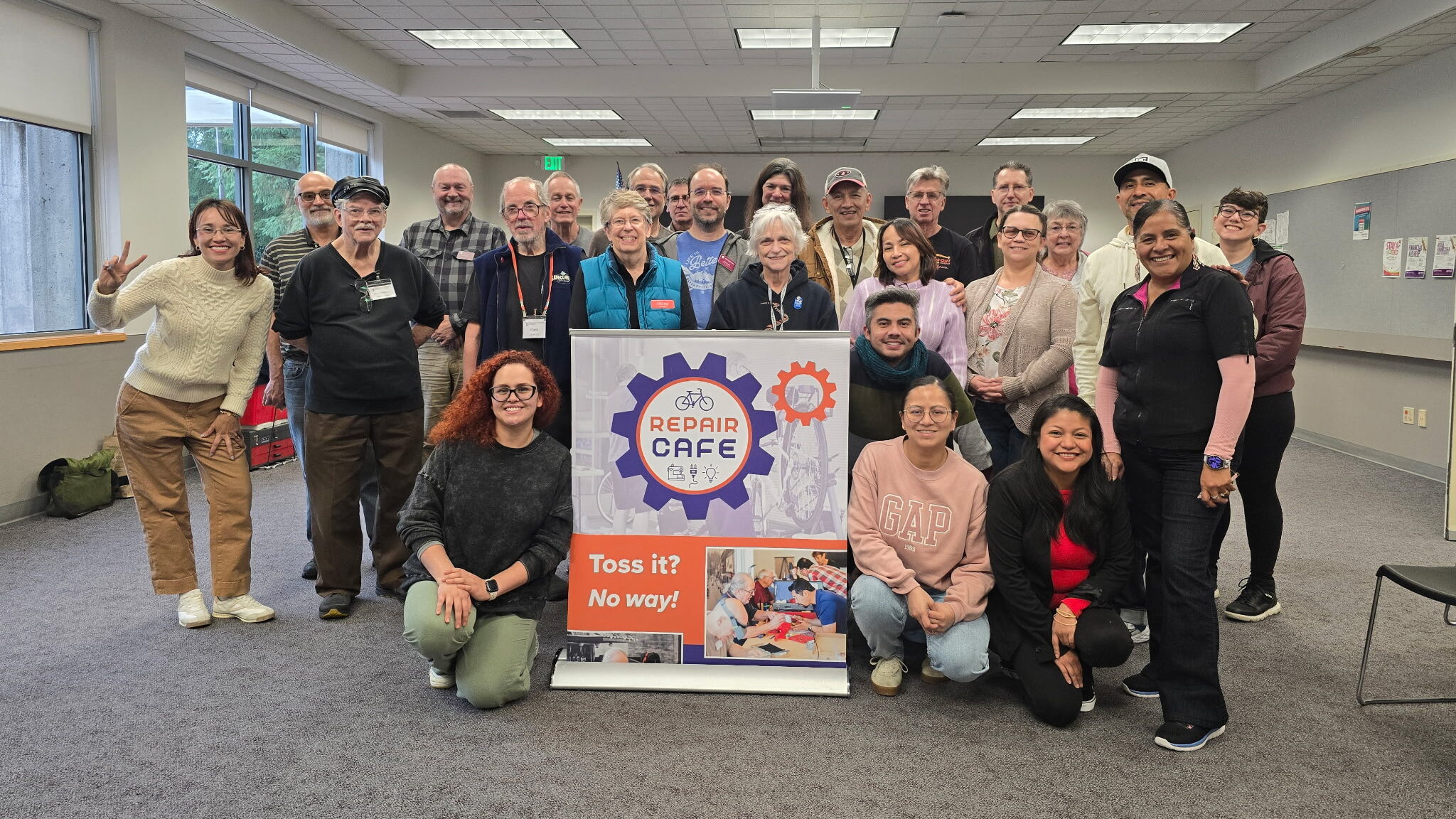 Group of smiling volunteers surround a Repair Cafe sign inside a meeting room.