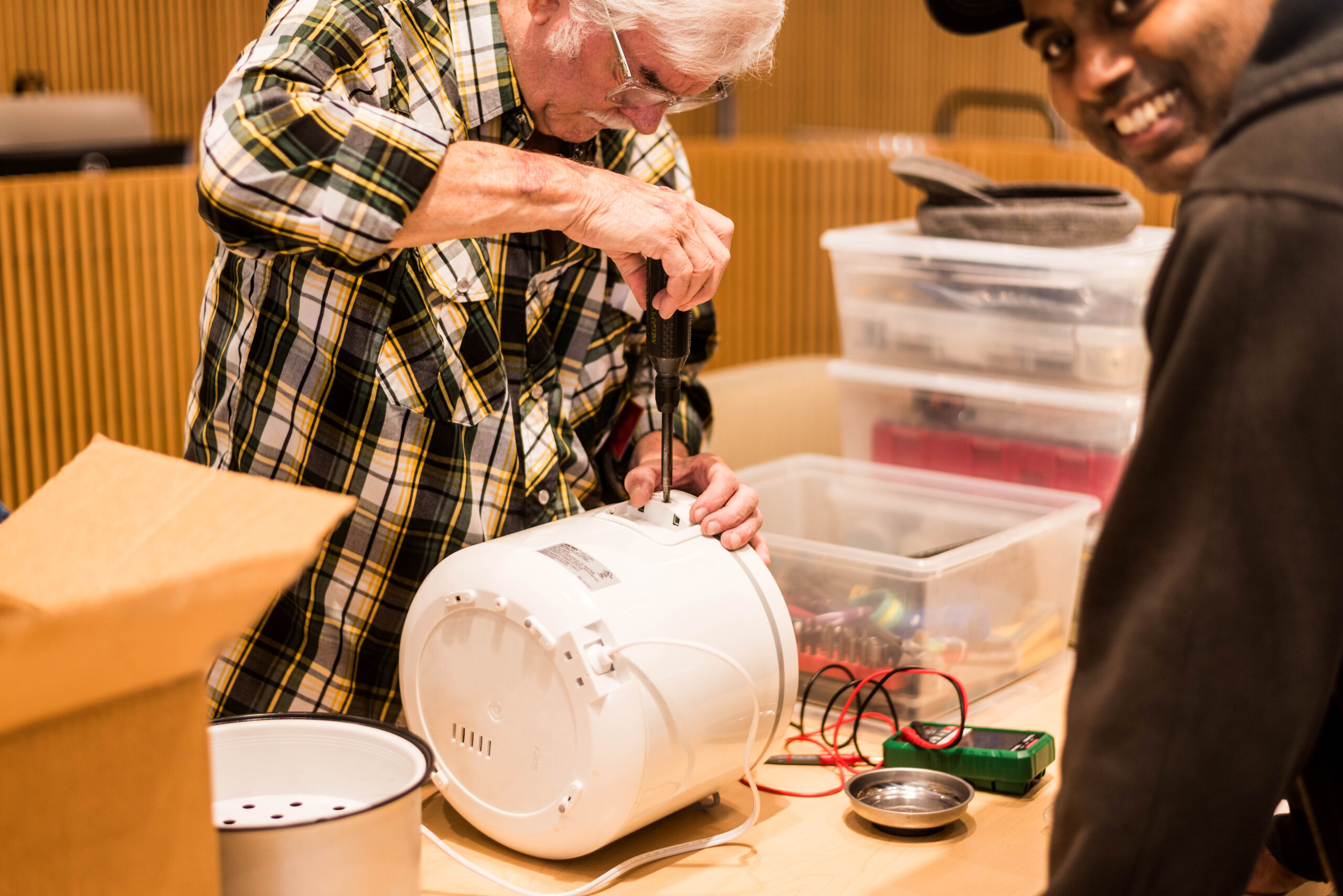 A man is working to repair a rice cooker while the owner smiles in the foreground.
