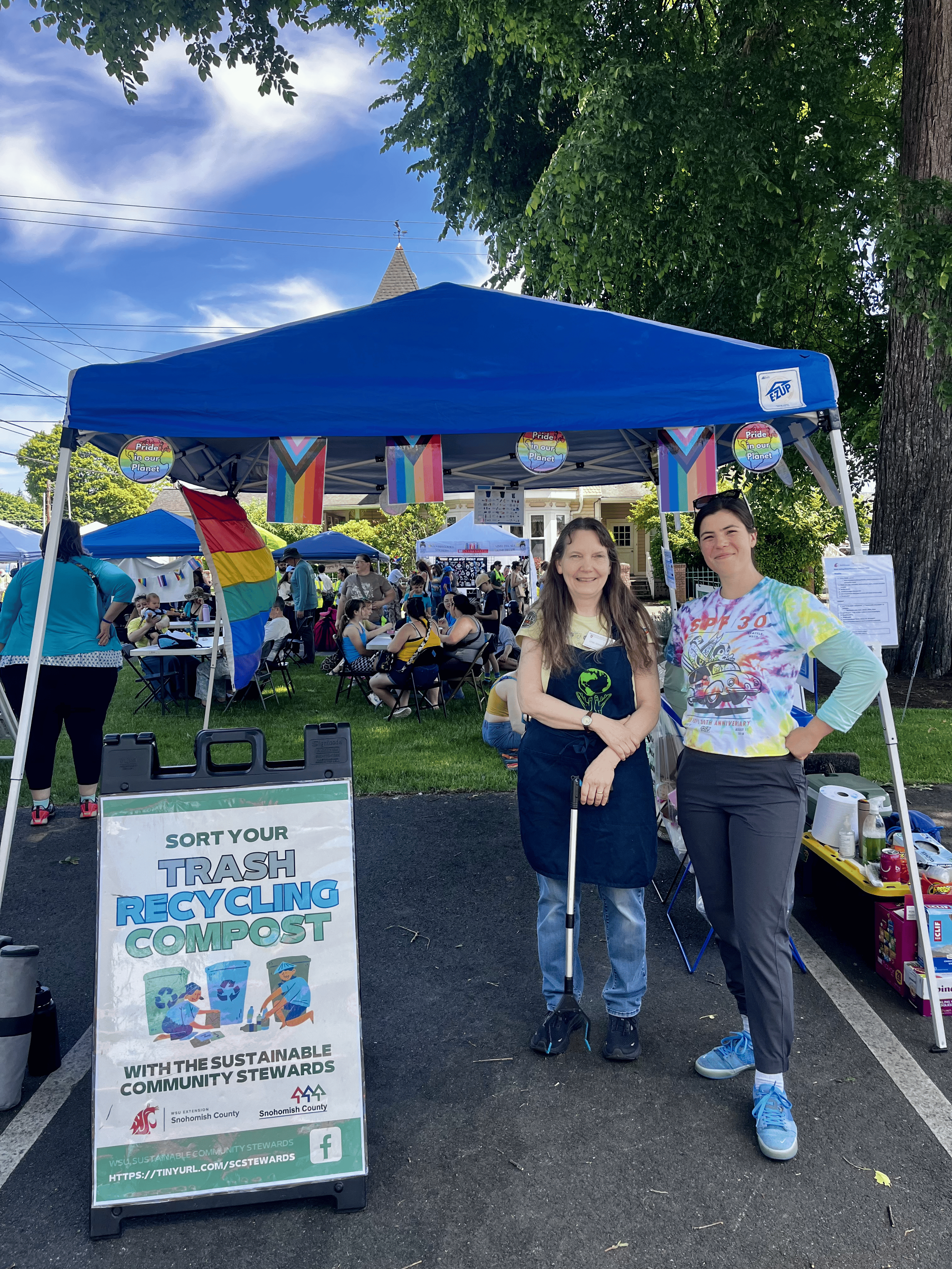 Two female volunteers stand in front of a waste station holding trash grabbers.