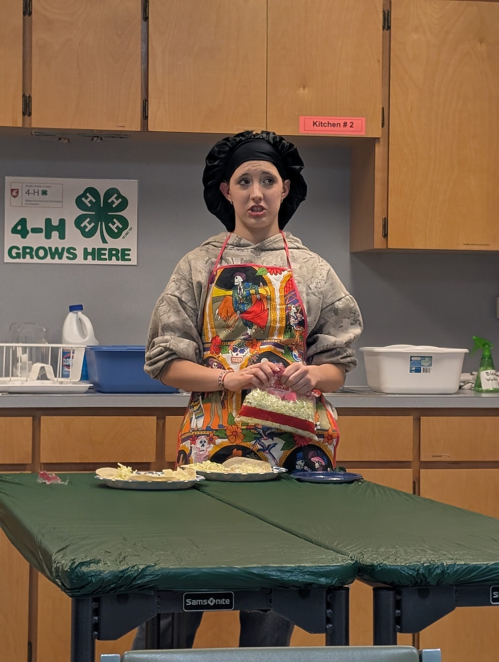 Person in apron and chef’s hat preparing food in a kitchen with a 4-H program sign.