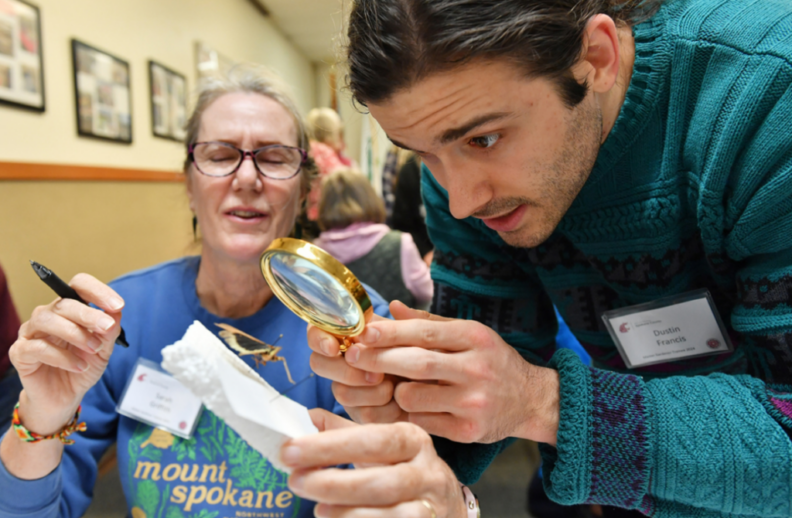 Master Gardener holds insect specimen while another Master Gardener examines with a hand lens.