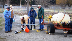 Group of men outdoors testing spraying equipment near a trailer with a large tank.