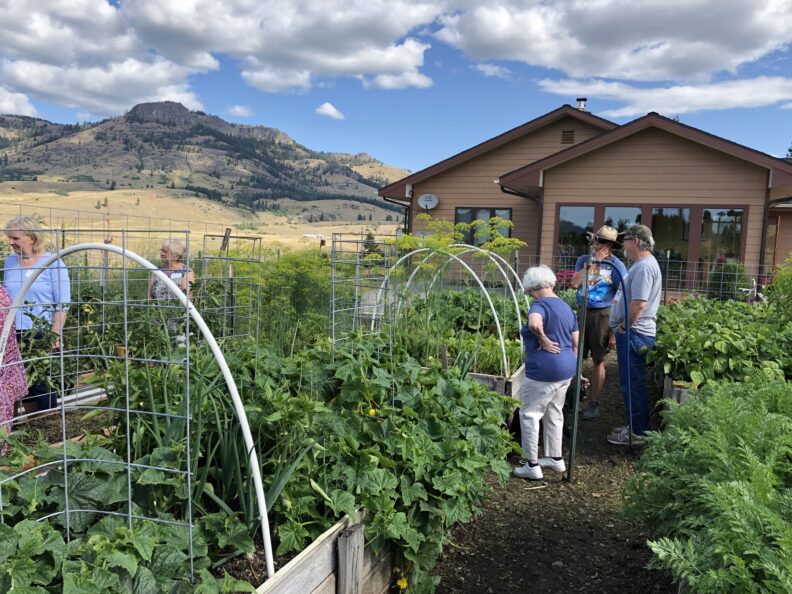 Group of people touring a lush garden with raised beds near a house, with hills and blue sky in the background.