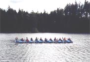 Group of people paddling a long boat on a lake, with trees along the shoreline.