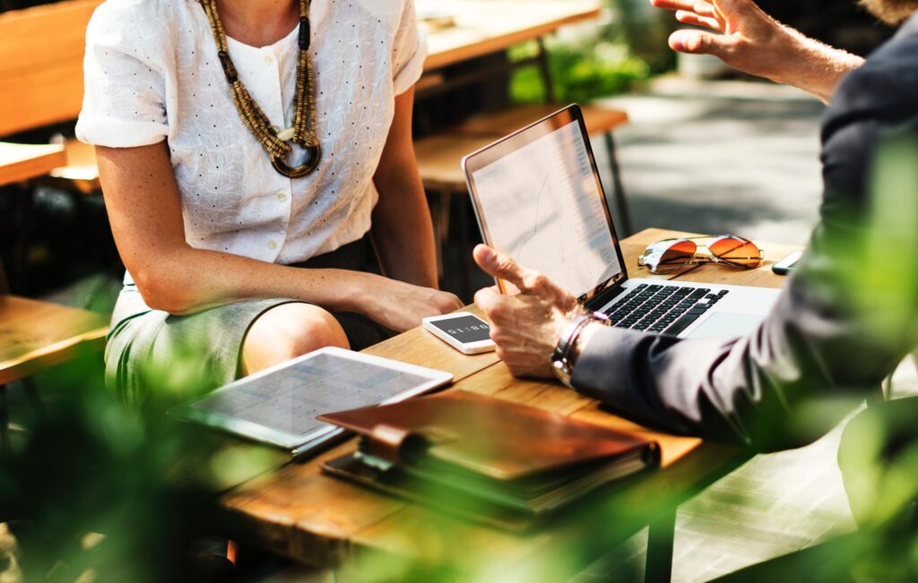 Two people having a discussion outdoors at a table with a laptop, tablet, and smartphone.