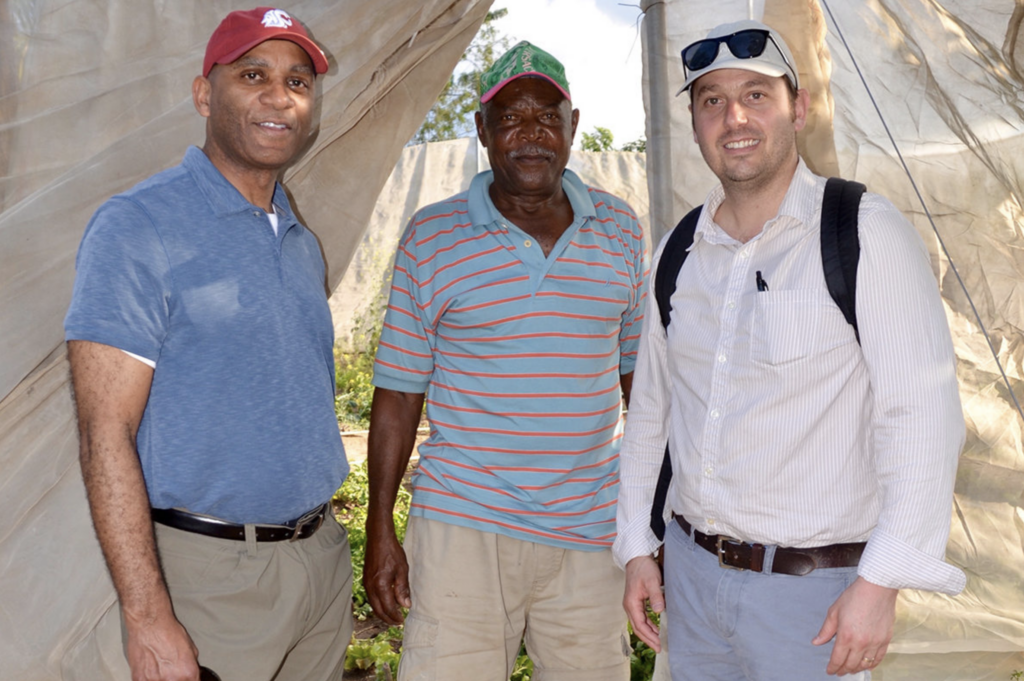 Three men standing inside a greenhouse structure, smiling at the camera on a sunny day.