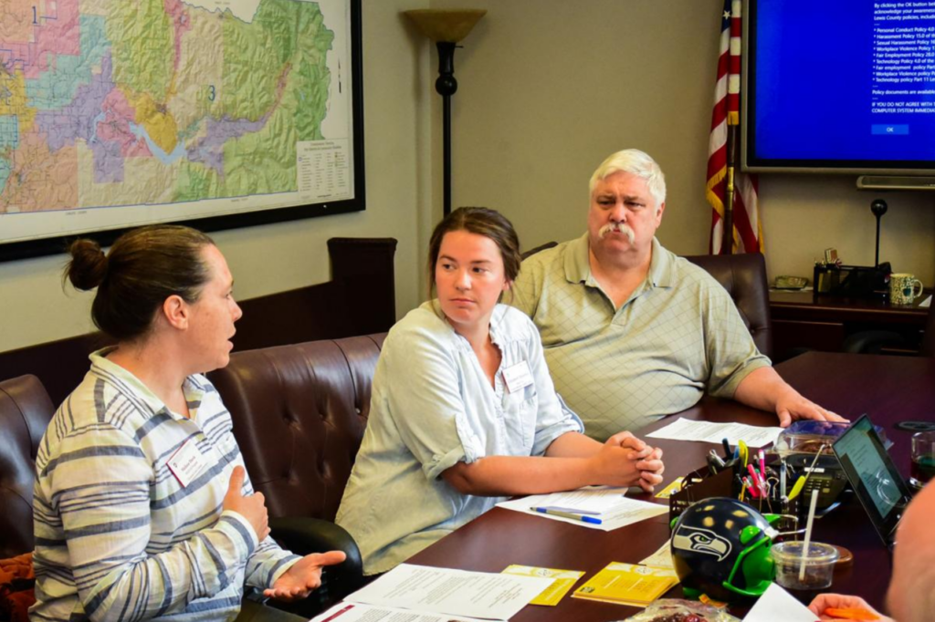 Three people in a meeting room discussing documents at a conference table, with maps and a screen in the background.