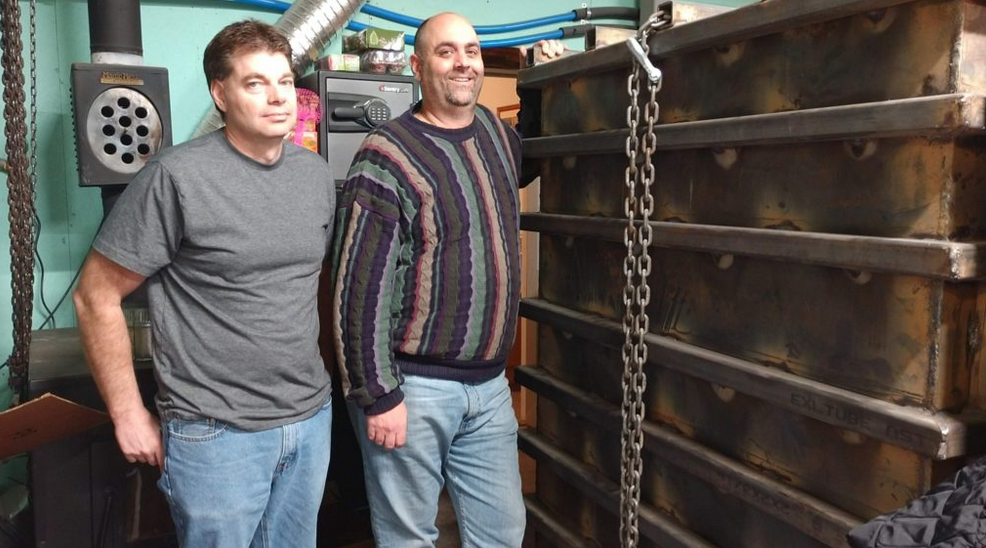 Two people standing beside a large metal industrial structure in a workshop or mechanical room.