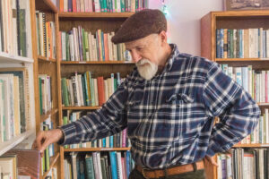 Older oerson in a plaid shirt and cap browsing books on a shelf in a cozy library or bookstore.