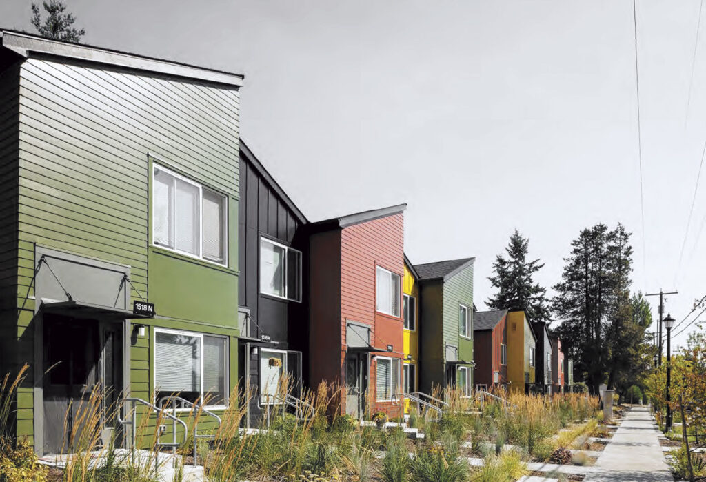 Row of modern, colorful townhouses with tall grass landscaping along a quiet sidewalk.